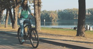 Beautiful summertime mood shot of young woman or girl riding bicycle through promenade, in stylish outift, pedalling next to trees in sun light, 4K video shooting by handheld gimbal