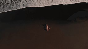 4K aerial top view flying video of young woman in bikini on the beach with black volcanic sand. Bali island.
