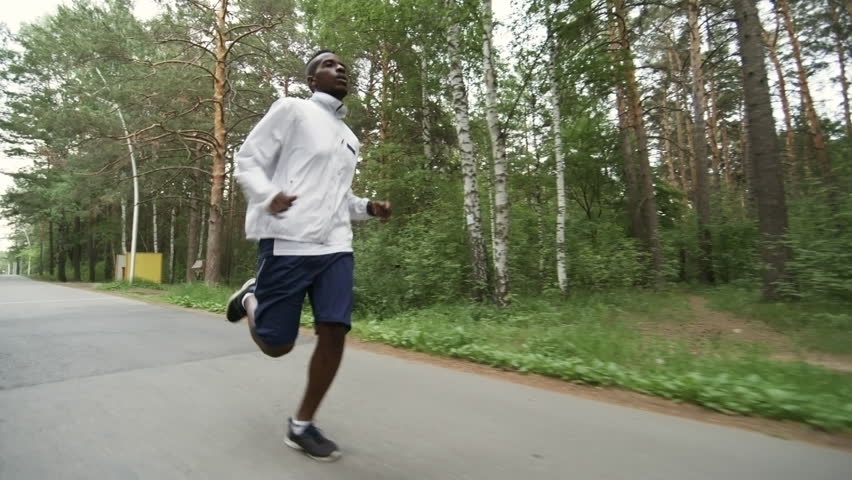 Low angle dolly shot of black man in windbreaker and short running along forest road on chilly morning