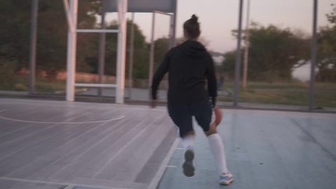 Backside footage of a young girl basketball player training and exercising outdoors on the local court. Dribbling with the ball, bouncing and make a shot