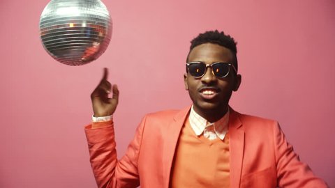 Portrait shot with pink background: cool black man in stylish jacket and sunglasses moving his finger in air and talking to camera while dancing and spinning disco ball