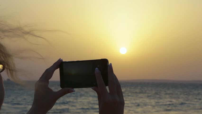 Happy european woman taking photo of sunrise or sunset sea using digital camera of smartphone during summer vacation. Closeup real time video of female hands making pictures of morning landscape.