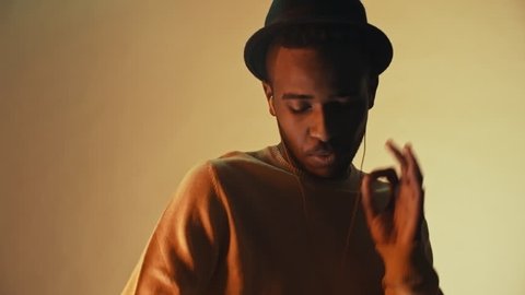 Low-key portrait shot of stylish black man in hat listening to music in earphones and rapping before camera in studio with yellow backdrop Video Stok