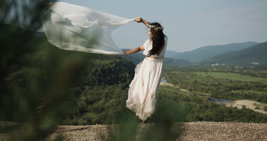 Wind blows woman's dress and veil while she stands before beautiful mo...