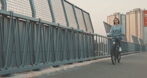 Young woman riding bicycle in city bridge on background buildings and sun. Gir bike rider drink coffee on background sky. Urban street scene . 4K video shooting by handheld gimbal