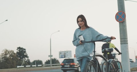 Young woman walking on city streets with bicycle and drinking coffee to go, sunshine, 4K video shooting by handheld gimbal