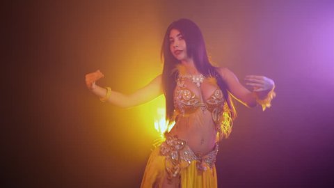Alluring sexy traditional oriental belly dancer girl dancing on yellow neon smoke background. Woman in exotic costume with feathers sexually moves her semi-nude body