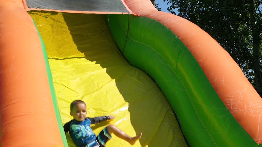 A slow motion shot of kids playing on a blow up bouce house water slide on a hot summer day