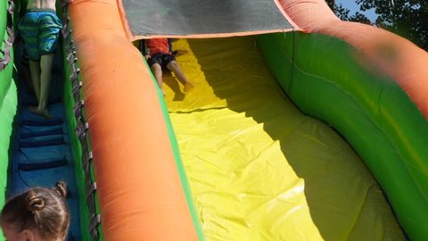 A slow motion shot of kids playing on a blow up bouce house water slide on a hot summer day
