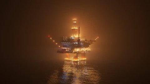 Big oil rig working on an open ocean during a foggy night. Countless lights illuminate the mist and create a reflection in the water waves. Symbol of natural environment degradation and pollution.
