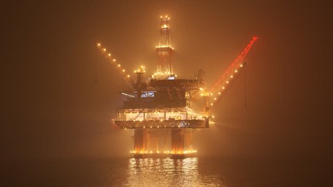 Big oil rig working on an open ocean during a foggy night. Countless lights illuminate the mist and create a reflection in the water waves. Symbol of natural environment degradation and pollution.
