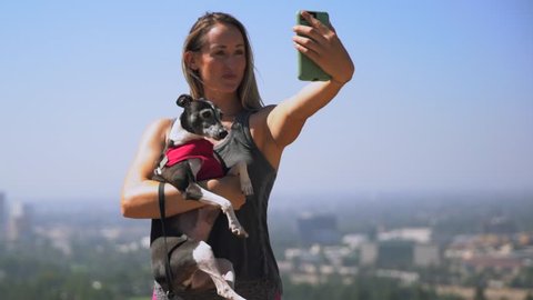 This real time video shows a young fit woman in her workout clothes taking scenic selfies on her cellphone with her italian greyhound dog.