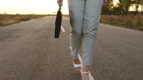 woman walks along asphalt, outside city in trousers and jacket and in white high heeled shoes, business woman carries black briefcase in her hand