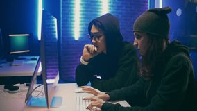 Team of hackers - a young girl and a man hacking a social network sitting at a table behind a large monitor with low lighting. 4k