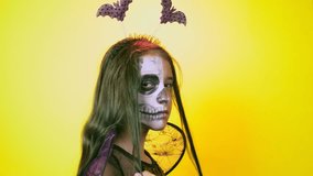 Halloween, girl with make-up skeleton on half face, dressed as witch, posing on bright yellow background. 4k, slow-motion, close-up