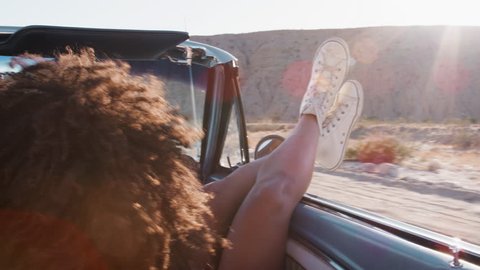 Young woman with legs sticking out of moving car, back view