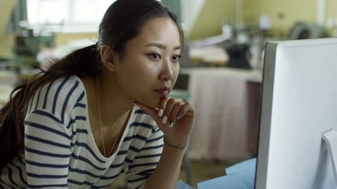 Medium shot of concentrated young Asian woman using desktop computer at work and thinking