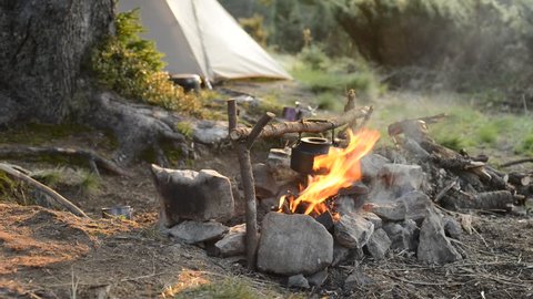 Old small kettle is heated on a bright bonfire on a green mountain meadow next to tent in the rays of rising sun - stunning morning during travel in the mountains.