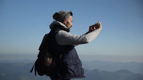 
Man in tourist equipment taking photos on the top of the mountain. Guy in grey beanie with backpack makes photo on phone on background of mountains.