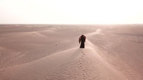 A single woman in abaya (United Arab Emirates traditional dress) walking on the dunes in the desert towards the sunrise bring light. Dubai, UAE.