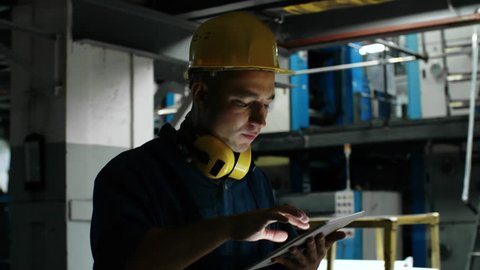 Medium follow shot of concentrated male worker in hard hat walking through factory and using tablet computer