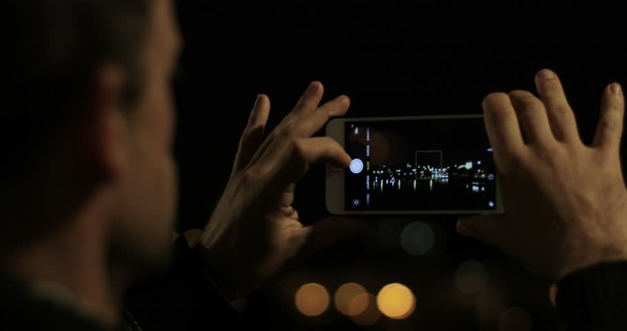 Man focusing making photo of night city lights on mobile phone close up no face black background. Male hands holding smartphone device picture screen of evening landscape water. Tourism concept

