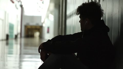High school teenager sitting in front of his locker alone in empty hallway.