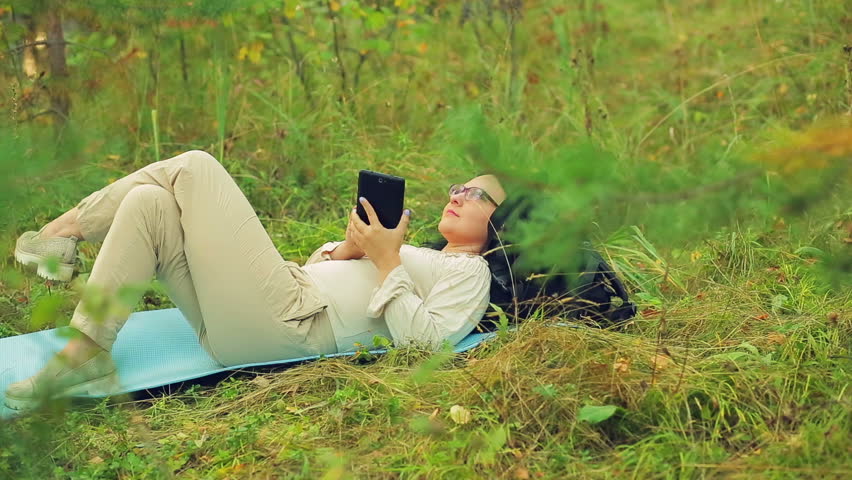 A young woman in glasses lies on the grass in the park and communicates in a messenger.