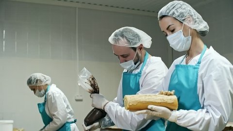 Tilt down shot of three confectioners in workwear making swiss rolls, woman spreading cream on cakes, and man using piping bag to decorate them with molten chocolate