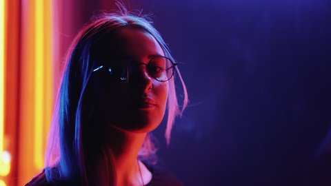 Millennial enigmatic pretty girl with unusual dyed hairstyle near glowing neon wall at night. Blue hair, golden sequins as freckles,nose piercing. Mysterious hipster teenager in glasses.