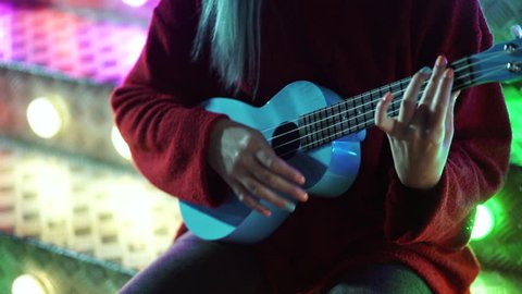Hands of young pretty girl playing on blue ukulele while sitting on glowing neon stairs in amusement park at night