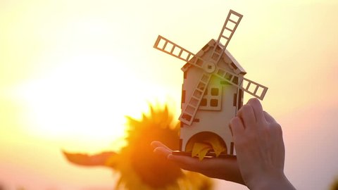 Girl hands holding a toy mill on a background of sunset and sunflowers. The mill slowly rotates. The concept of harvesting.