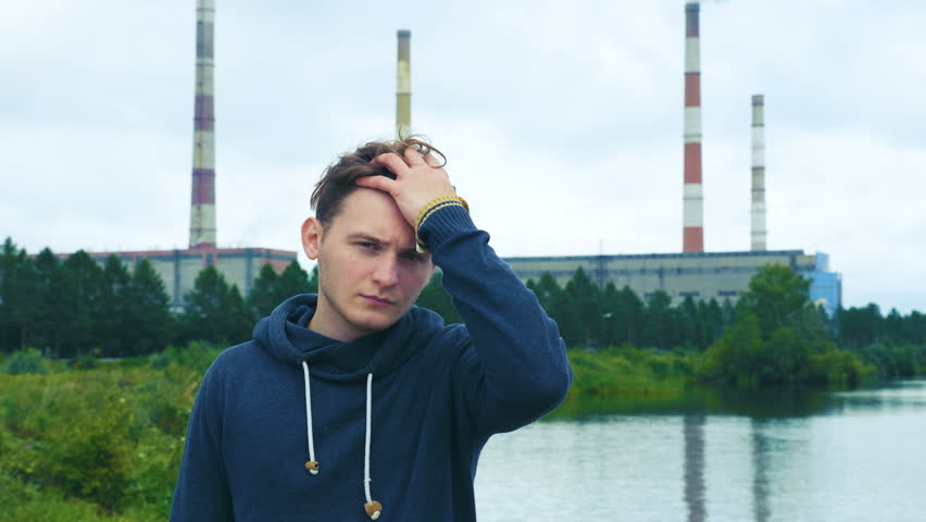 Portrait of a tired, sleepy, discontented young man standing in the background of the hydroelectric power station and the cloudy sky.