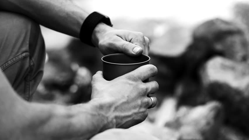 Man traveler hands holding cup of coffee near fire outdoors. Adventure, travel, tourism, camping concept. Hiker drinking tea from mug at camp. Coffee cooked over campfire on nature. Black and white.