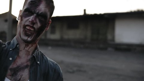 Portrait of a male zombie with bloody teeth and wounded face screaming and shouting. Halloween, filming, staging concept. Blurred abandoned town on the background