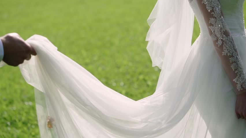 Bride and groom walking across park at sunny summer day. Holding hands, slowmotion