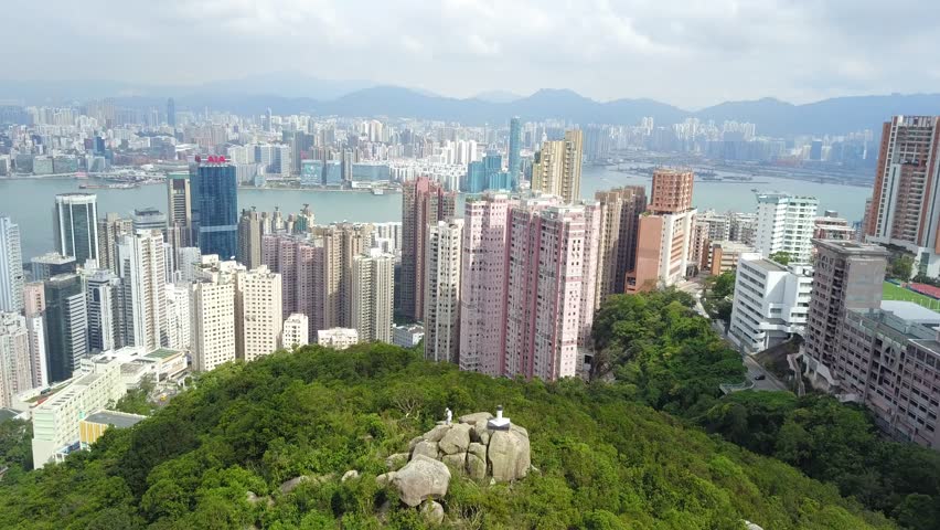 Aerial panorama of Hong Kong viewed from top of Braemar Hill Peak, with a city skyline of crowded skyscrapers by Victoria Harbour, residential towers on hillside & Kowloon downtown across the seaport