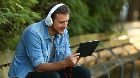 Portrait of a happy guy relaxing watching videos on line in a tablet sitting on a bench in a park