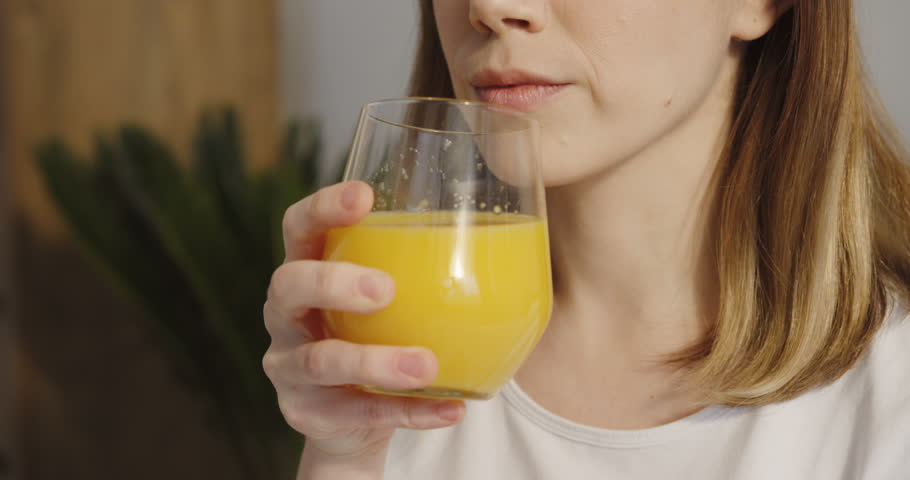 Close up of the young Caucasian pretty woman drinking orange juice from a glass and smiling to the camera.