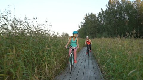 Two young women ride bicycles on a wooden ecological trail among the reeds