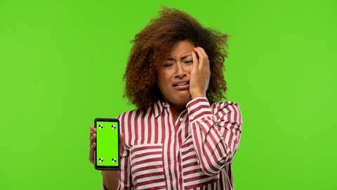 Afro american woman holding phone on green chroma desperate and sad, crying inconsolably, is pessimistic and melancholy