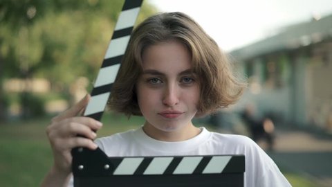 Serious young woman with short thick hair looking at camera. She is using clapperboard and starts laughing after that. Concept of filmmaking and acting. Slider slow motion portrait shot