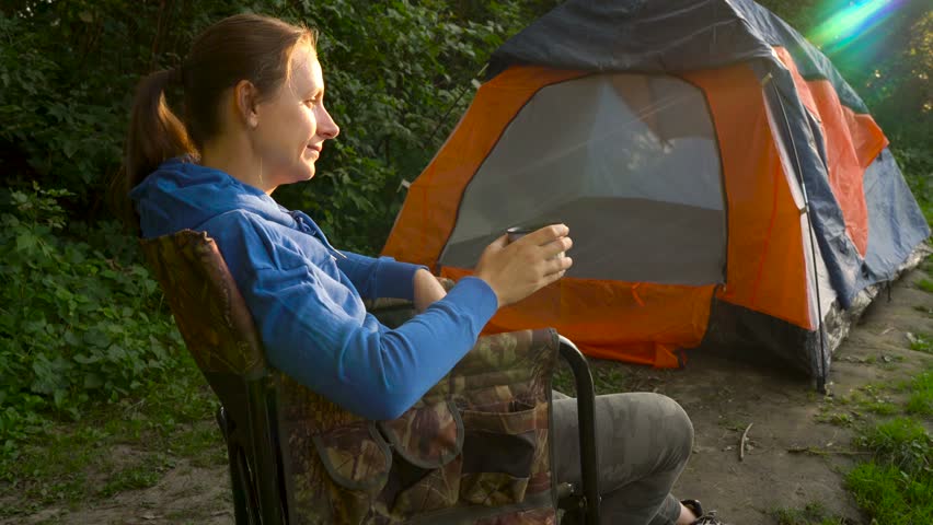 Woman is drinking tea in an armchair near a tent on the river bank