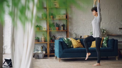 Active person is doing yoga at home practising balance exercises on one leg standing on floor alone. Beautiful loft style flat with furniture and plants is visible.