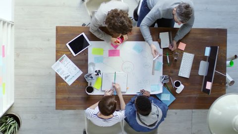 Top view of group of four multiethnic business people sitting at desk in office and using sticky notes to create mind map on large sheet of paper