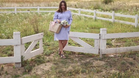 Beautiful pregnant woman in blue dress walking hear the farm. Knitted bag with bouquet of flowers. Background of tree and mountains. Slow motion