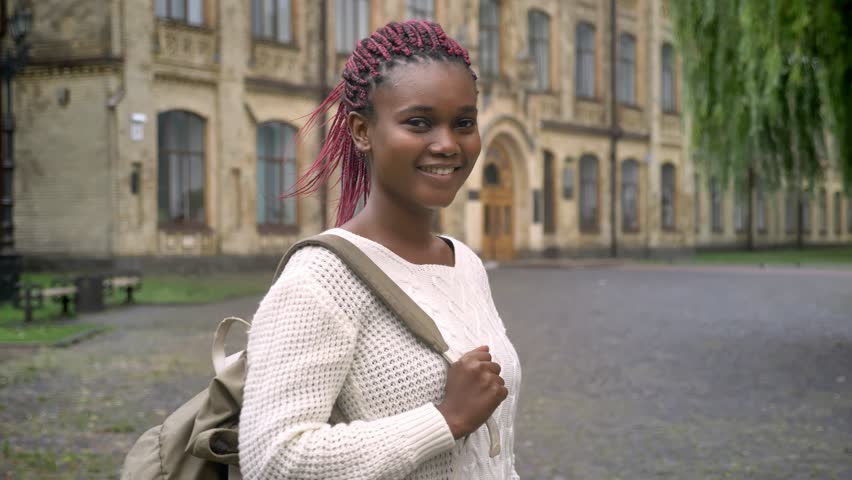 Young african american woman with pink dreadlocks holding backpack and smiling at camera, standing in park near university, happy