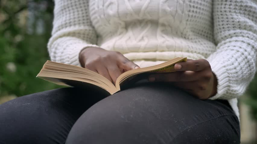 Moving close footage of young african american student reading book and sitting on bench in park near university