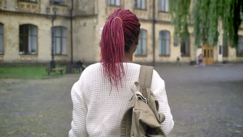 Moving shot of young african female student going to university, charming woman with backpack and pink dreadlocks