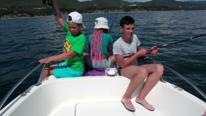 Three happy teen age kids sitting in boat with spinning tackles, have fishing excursion
